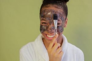 happy black teen applying exfoliating mask on face with applicator